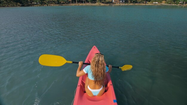 Mujer joven sexy en traje de baño azul balancea los pies en el agua en canoa rosa en el mar contra la parte trasera de las colinas