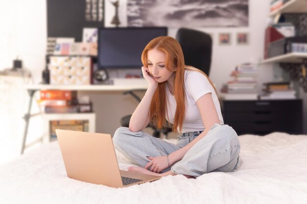 La mujer joven se sienta con la computadora portátil en la cama en casa