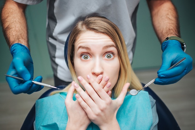 La mujer joven se sienta inchair en odontología. Ella tiene miedo y se tapa la boca con las manos. Cliente mira a la cámara con miedo en los ojos. Equipo de retención de dentista masculino para el tratamiento de los dientes.