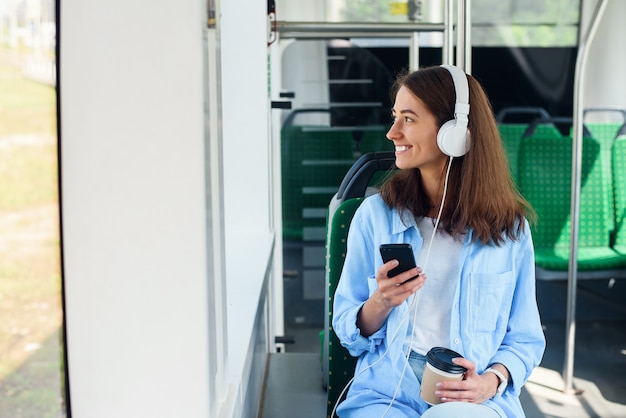 Una mujer joven se sienta en el moderno autobús de la ciudad, escucha música, bebe café y mira por la ventana.