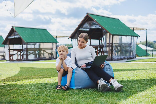 Una mujer joven se sienta en una silla blanda en el parque y trabaja en una computadora portátil mientras su hijo juega junto a ella Trabajando al aire libre con licencia de maternidad