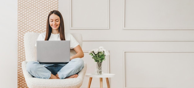 La mujer joven se sienta en una silla y trabaja con una computadora portátil en casa contra el fondo de una pared blanca