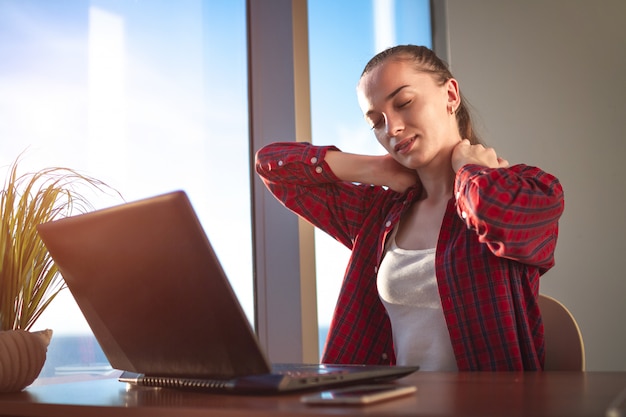 La mujer joven siente molestias en la columna cervical y masajea el cuello rígido en el trabajo.