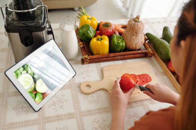 Mujer joven siguiendo la receta en la computadora de la tableta al cocinar la cena y cortar verduras frescas