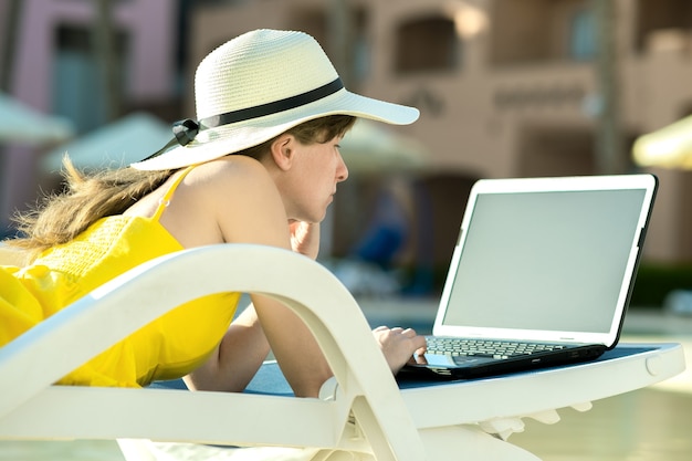 Mujer joven en silla de playa en la piscina trabajando en equipo portátil conectado a internet inalámbrico escribiendo texto en las teclas en el lugar de veraneo. Trabajo a distancia y trabajo independiente mientras viaja concepto.