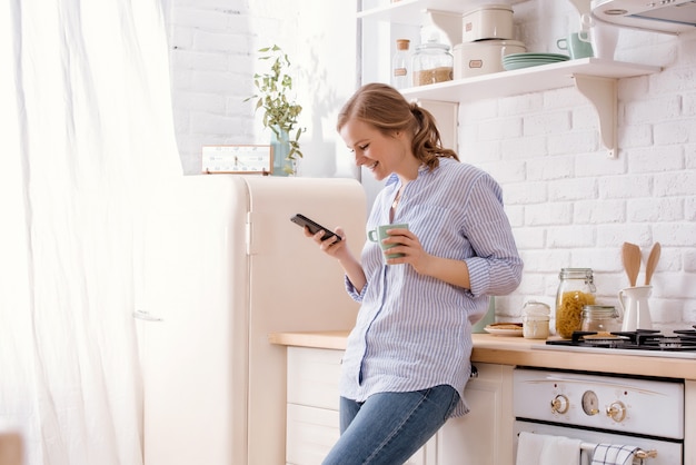 Mujer joven con smartphone apoyado en la mesa de la cocina con taza de café y organizador en un hogar moderno. Mujer sonriente que lee el mensaje de teléfono. Chica morena feliz escribiendo un mensaje de texto.