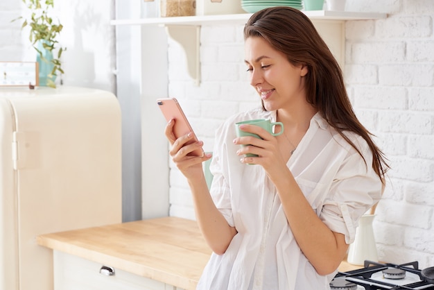 Mujer joven con smartphone apoyado en la mesa de la cocina con taza de café y organizador en un hogar moderno.