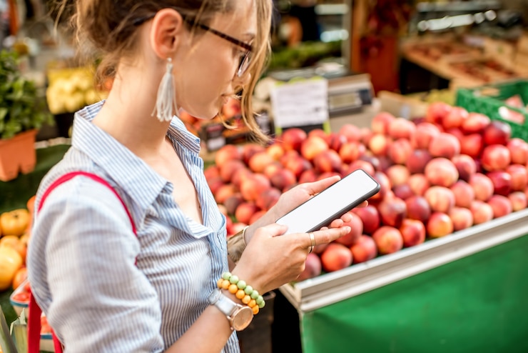 Foto mujer joven con smartphone de pie con bolsa frente al mercado de alimentos en francia