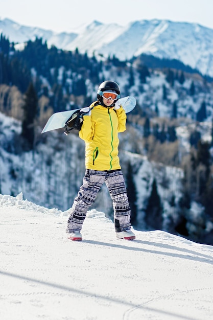 Mujer joven snowboarder caminando sobre la pista de esquí sosteniendo su tabla de snowboard sobre sus hombros