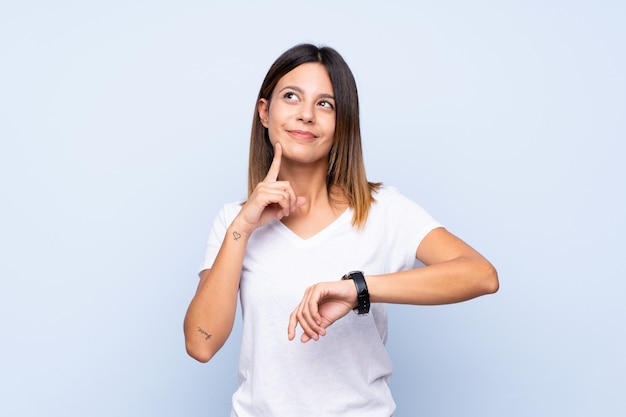 Mujer joven sobre azul aislado con reloj de pulsera y pensando en una idea