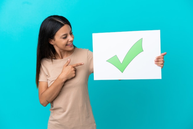 Mujer joven sobre fondo aislado sosteniendo un cartel con el icono de marca de verificación verde de texto y apuntando