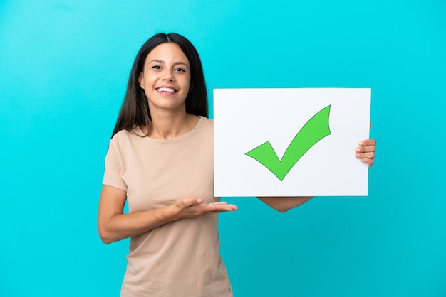 Mujer joven sobre fondo aislado sosteniendo un cartel con el icono de marca de verificación verde de texto y apuntando