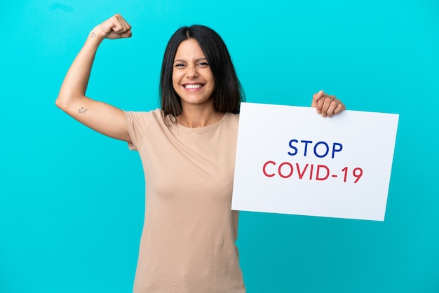 Mujer joven sobre fondo aislado sosteniendo un cartel con el texto Stop Covid 19 y haciendo un gesto fuerte