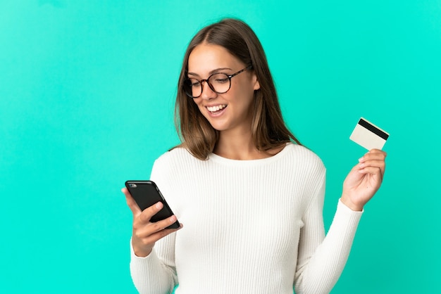 Mujer joven sobre fondo azul aislado comprando con el móvil con tarjeta de crédito