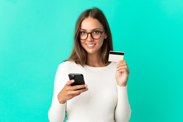 Mujer joven sobre fondo azul aislado comprando con el móvil con tarjeta de crédito