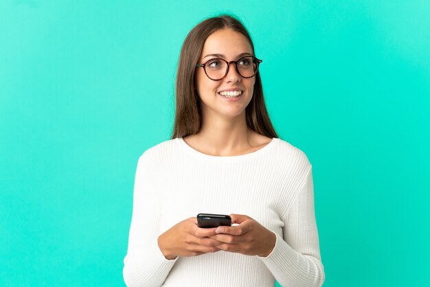 Foto mujer joven sobre fondo azul aislado mediante teléfono móvil y mirando hacia arriba