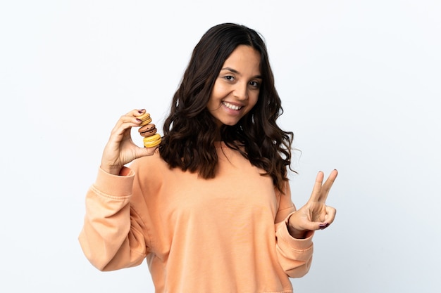 Mujer joven sobre fondo blanco aislado sosteniendo coloridos macarons franceses y mostrando el signo de la victoria