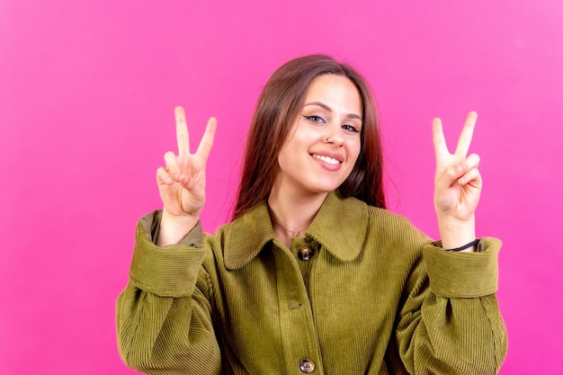 Mujer joven sobre fondo rosa sonriendo y mostrando el signo de la victoria vistiendo una cazadora verde