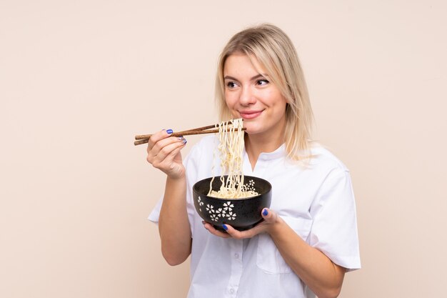 Mujer joven sobre pared aislada sosteniendo un tazón de fideos con palillos