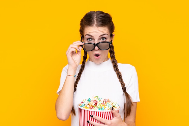 Mujer joven sobre pared amarilla aislada sorprendida con gafas 3d y sosteniendo un gran cubo de palomitas de maíz