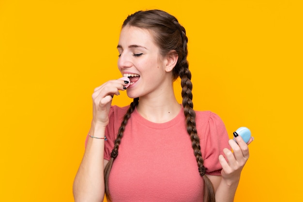 Mujer joven sobre pared amarilla aislada sosteniendo coloridos macarons franceses y comiendo