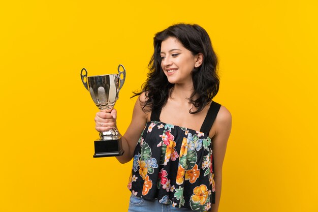 Mujer joven sobre pared amarilla aislada sosteniendo un trofeo