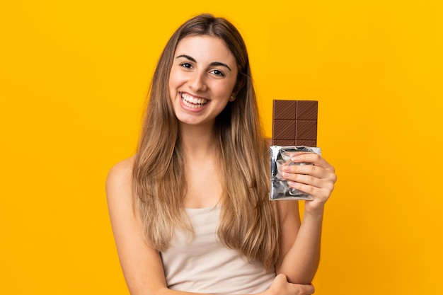 Mujer joven sobre pared amarilla aislada tomando una tableta de chocolate y feliz