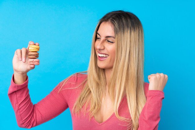 Mujer joven sobre pared azul aislada con coloridos macarons franceses y celebrando una victoria