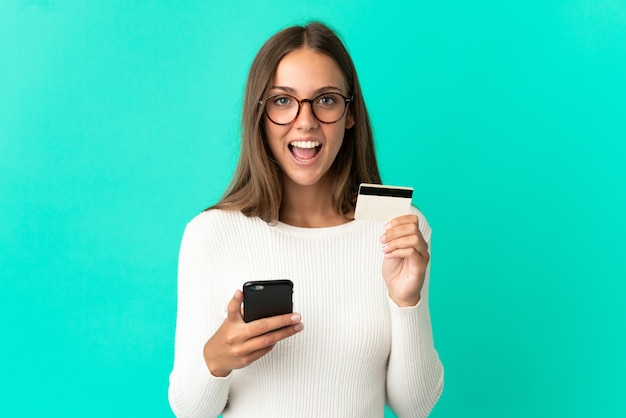 Mujer joven sobre pared azul aislada comprando con el móvil y sosteniendo una tarjeta de crédito con expresión de sorpresa