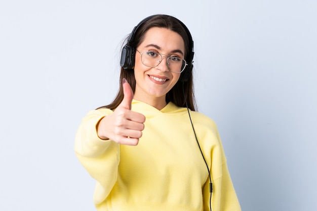 Mujer joven sobre pared azul aislada escuchando música y con el pulgar arriba