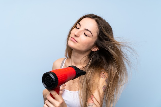 Mujer joven sobre pared azul aislada con secador de pelo