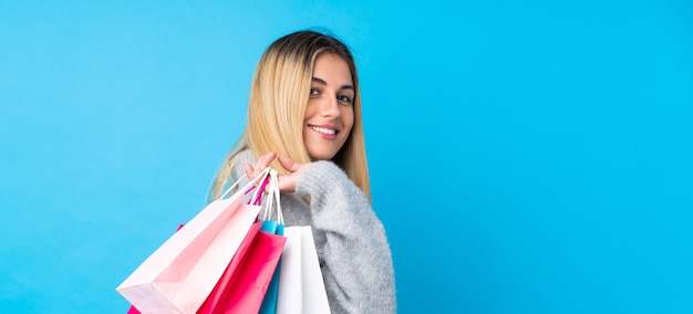 Mujer joven sobre pared azul aislada sosteniendo bolsas de compras y sonriendo