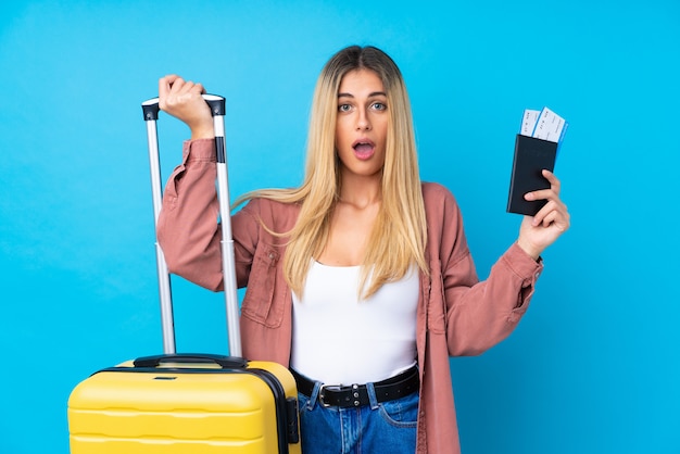 Mujer joven sobre pared azul aislada en vacaciones con maleta y pasaporte y sorprendida