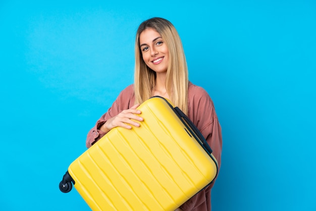 Mujer joven sobre pared azul aislada en vacaciones con maleta de viaje