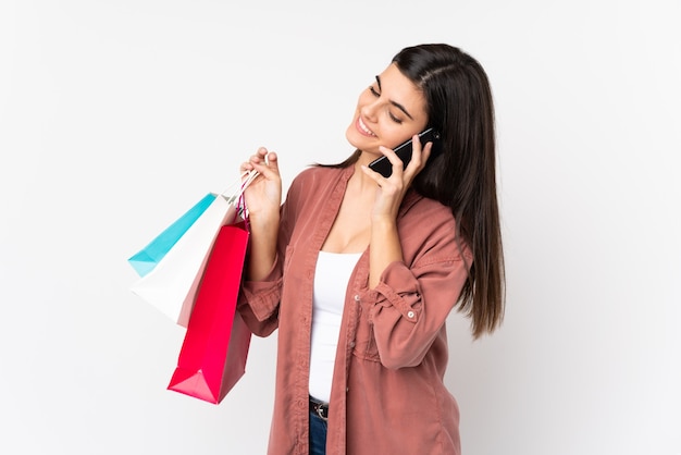 Mujer joven sobre pared blanca aislada sosteniendo bolsas de compras y llamando a un amigo con su teléfono celular