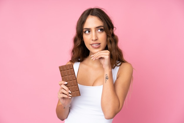 Foto mujer joven sobre pared rosa aislada tomando una tableta de chocolate y teniendo dudas