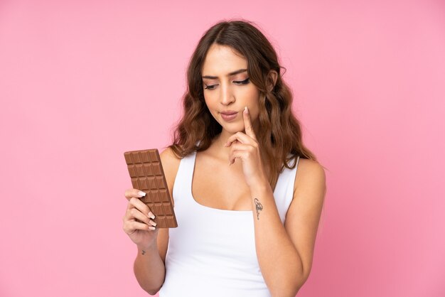 Mujer joven sobre pared rosa aislada tomando una tableta de chocolate y teniendo dudas