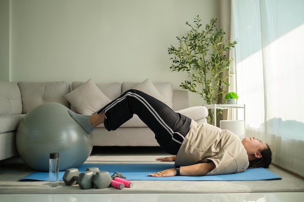 Mujer joven con sobrepeso haciendo ejercicio con una pelota de fitness en la alfombra en casa Estilo de vida saludable