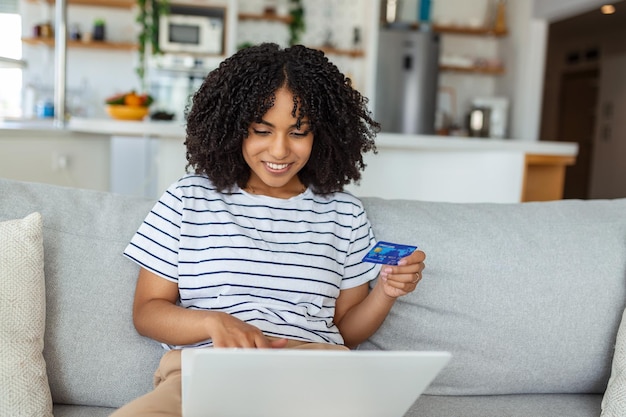 Mujer joven en el sofá comprando en línea con tarjeta de débito Hermosa mujer africana usando una computadora portátil para comprar en línea en casa