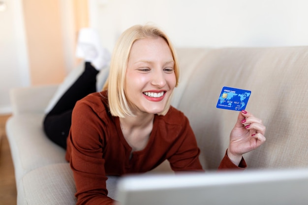 Mujer joven en el sofá Compras en línea con tarjeta de débito Hermosa chica usando una computadora portátil para comprar en línea en casa