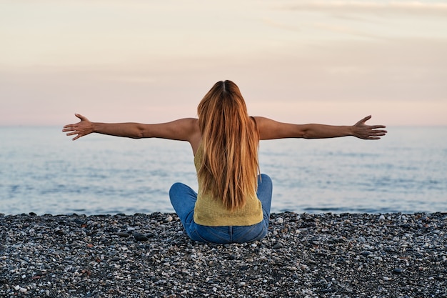 Mujer joven sola sentada con los brazos abiertos en la arena de la playa al atardecer. Concepto de relajación, meditación y libertad.