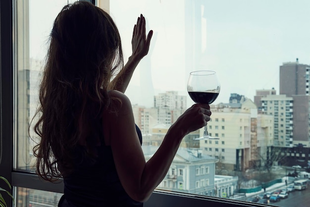 Mujer joven solitaria en casa bebiendo alcohol Alcoholismo femenino sola mujer hermosa de lujo en vestido negro con vino de pie cerca de la ventana mirando a un lado
