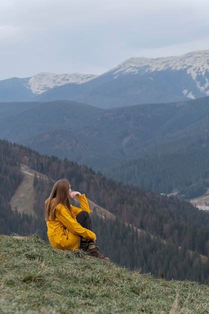 Mujer joven solitaria en las montañas disfrutando del aire limpio y la naturaleza Chica de chaqueta amarilla se sienta en la ladera de la montaña Vertical