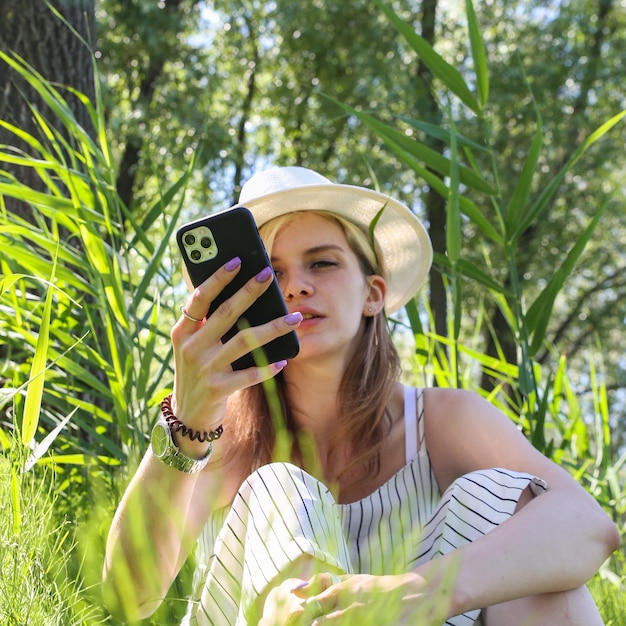 una mujer joven con un sombrero blanco está sentada en la hierba con un teléfono en la mano