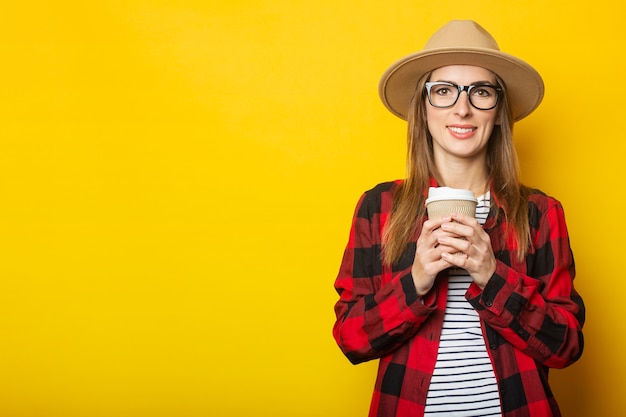 Mujer joven con sombrero y camisa a cuadros sosteniendo una taza de papel con café en amarillo