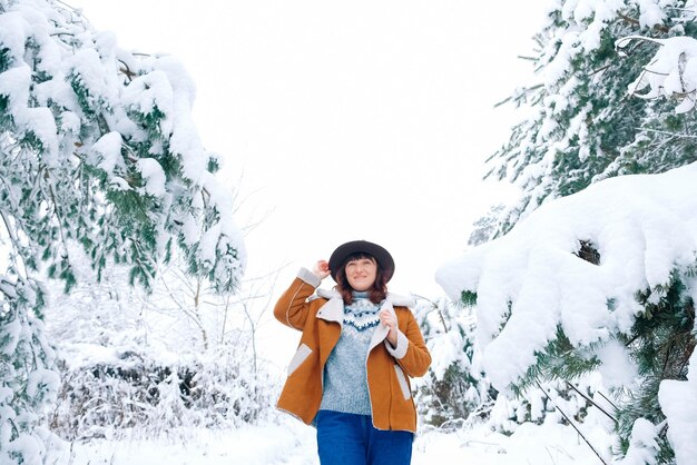 Mujer joven con sombrero y chaqueta abrigada de pie entre árboles nevados y disfrutando de la primera nieve