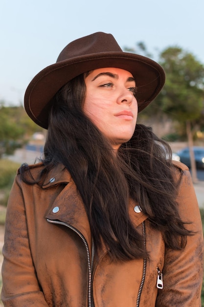 Mujer joven con sombrero y chaqueta de cuero marrón mirando a la distancia en un parque natural