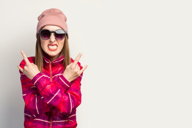 Foto mujer joven con sombrero y gafas y una chaqueta deportiva rosa hace un gesto de cabra de rock and roll.