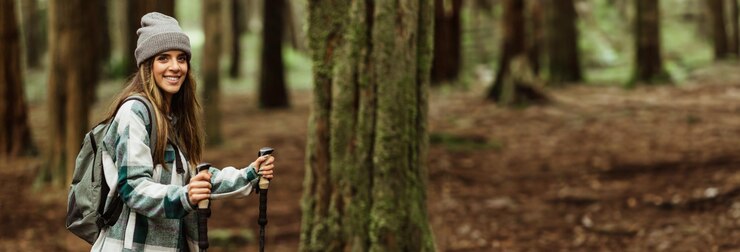Una mujer joven con un sombrero gris y una camisa a cuadros camina por un bosque con una mochila y
