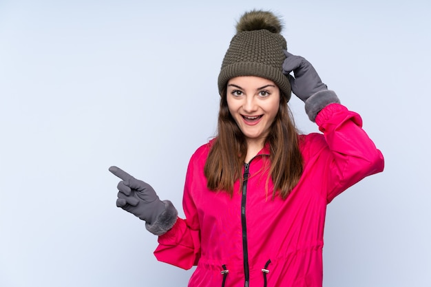Mujer joven con sombrero de invierno aislado en la pared azul sorprendido y apuntando con el dedo al lado